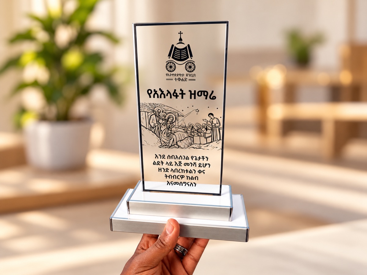 A hand holding up a clear printed acrylic award with Amharic text and a line illustration, on a brushed aluminium base, in a bright room with plants
