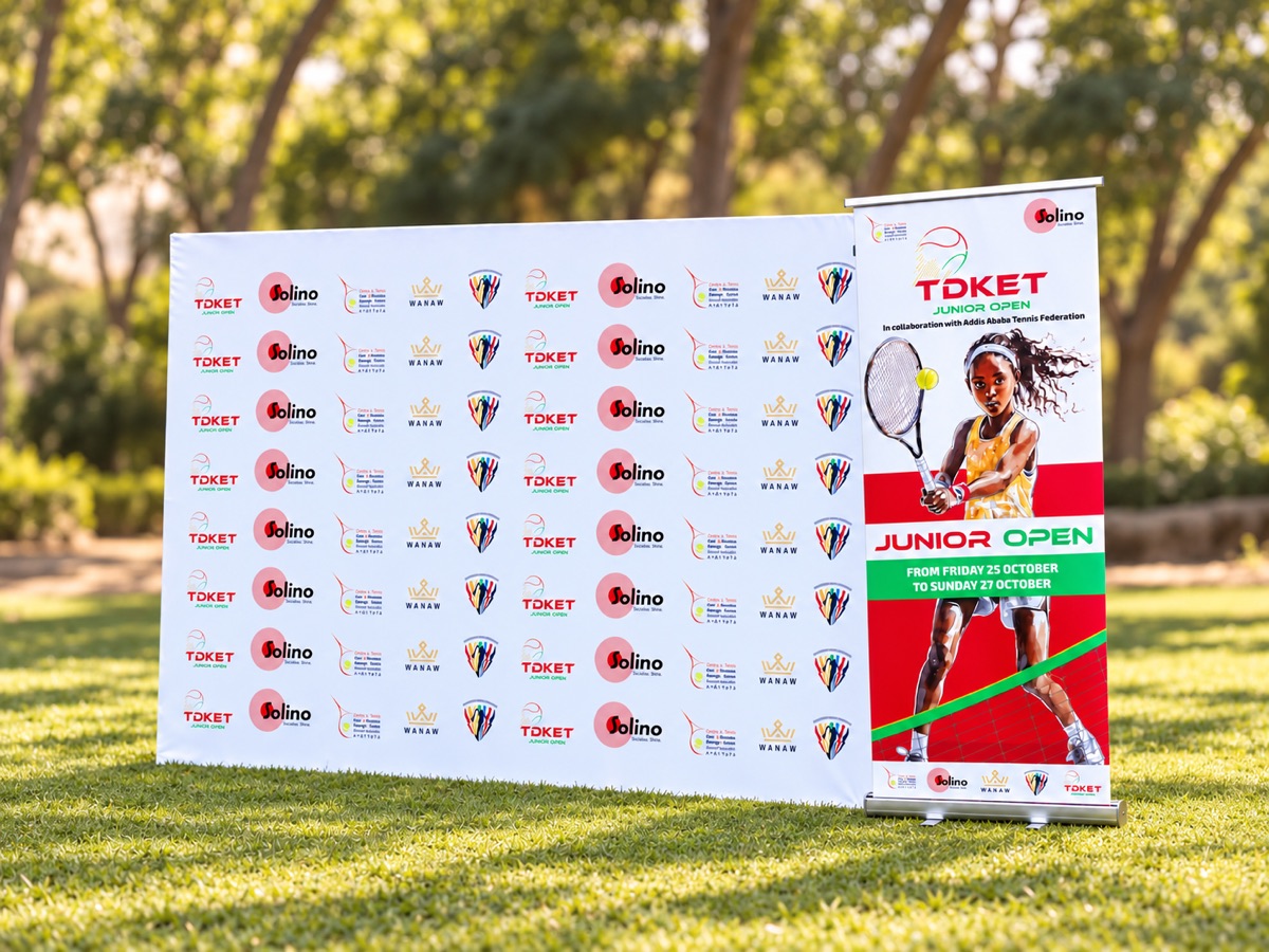 A wide step-and-repeat backdrop printed with rows of sponsor logos, beside a roll-up banner showing a young tennis player, set up on a lawn under trees