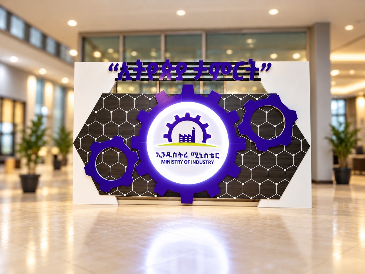 An event backdrop shaped like interlocking purple gears on a hexagon-pattern panel, with a lit circular logo in the centre and raised Amharic lettering along the top, in a bright lobby