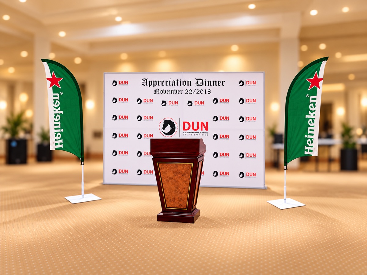 A step-and-repeat backdrop for an appreciation dinner with a wooden podium in front and a green feather flag on each side, in an empty carpeted hall