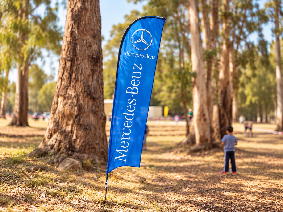 A tall blue feather flag with white lettering staked in the ground among eucalyptus trees in a sunny park, with a child in the background